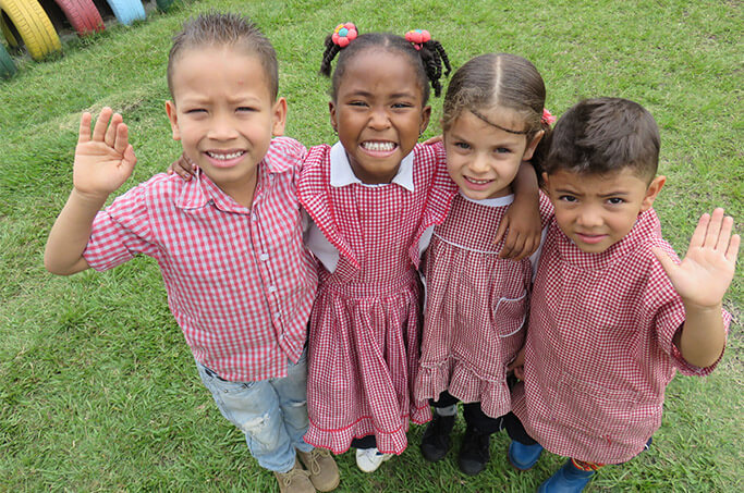 Cuatro niños abrazados, sonrientes y saludando a la cámara.